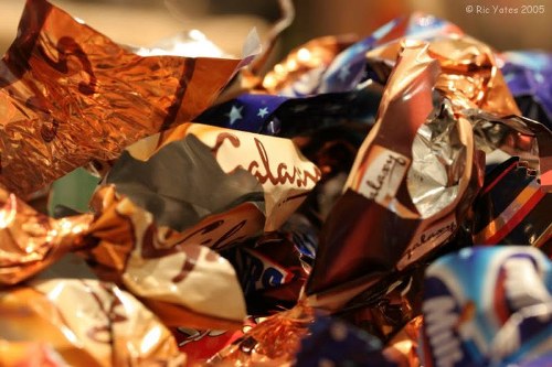 A close-up view of a pile of crumpled chocolate wrappers and foil packaging materials, predominantly in metallic copper, silver, and vibrant multicolour designs, scattered on a surface. The wrappers display various textures, with some shiny and reflective, while others appear matte or textured from being crushed. The items are overlapping, creating a chaotic arrangement typical of discarded confectionery packaging after consumption. In the background, there is a blurred surface that may be part of a rubbish collection or disposal area, suggesting the context of waste removal. This detailed visual accurately depicts waste materials related to household rubbish, aligning with services provided by Commercial Waste Removal Bermondsey in managing waste collections, including packaging cleanup, within the Bermondsey area, postcode SE1. The image emphasizes the importance of proper disposal and waste management for discarded food wrappers and packaging materials.