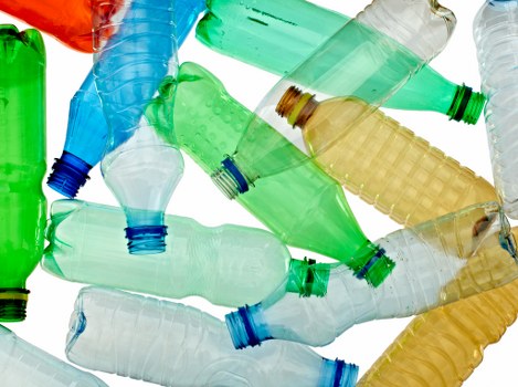 A collection of discarded plastic bottles, primarily made of PET with various colours including green, clear, blue, and light brown. The bottles are crumpled and stacked randomly on a white background, with some lying flat and others leaning or overlapping. The bottle caps are predominantly blue and green, with a few missing or loosened. The plastic surfaces display different textures, from smooth to slightly ridged, and some bottles show visible scuff marks or creases from use or compression. The arrangement of the bottles indicates they are waste materials awaiting collection or recycling, fitting within the context of rubbish removal services. The overall image highlights the typical type of domestic or commercial plastic waste that companies like Commercial Waste Removal Bermondsey manage, emphasizing the importance of proper disposal and recycling of plastics in the Bermondsey area.