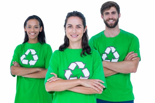 Three waste management professionals stand outdoors in front of a building, all wearing bright green T-shirts featuring a white recycling symbol on the front, indicating their association with rubbish removal services. The woman in the centre has shoulder-length dark hair and is smiling directly at the camera with her arms crossed. To her left, a woman with long dark hair also smiling, has her arms crossed, while to her right, a man with a beard and short dark hair, smiling broadly, likewise has his arms crossed. Behind them, the background shows a brick or concrete structure, suggesting an urban or industrial setting, with natural daylight illuminating the scene. The individuals are dressed practically for manual work, and their confident expressions imply readiness to assist with waste clearance tasks in the Bermondsey area or nearby parts of London. The image projects a professional and approachable impression of a commercial rubbish removal team, emphasizing their role in waste management and recycling within the local community, aligning with services offered by Commercial Waste Removal Bermondsey and related to the rubbish service category.