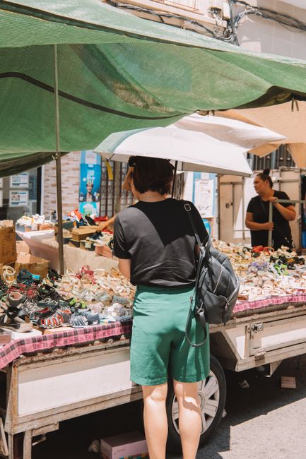A woman with dark hair, wearing a dark grey t-shirt and mint green shorts, stands with her back to the camera, browsing a market stall that is set up on the back of a small truck. The stall displays a